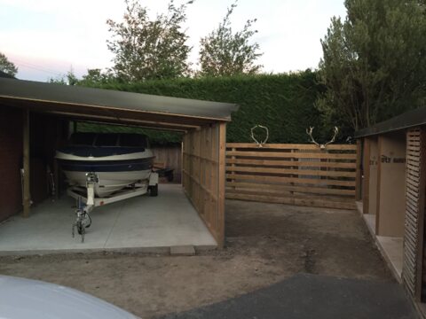 The carport seen from the drive, with a horizontal slat fence between it and the timber-framed firewood bays, and a clipped hedge behind