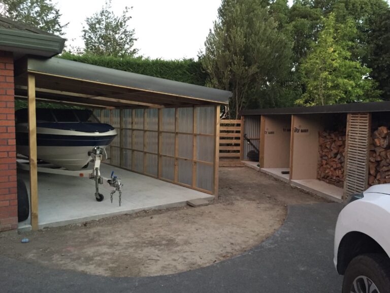 Timber-framed carport with translucent roof and wall sheeting sheltering a boat on a trailer, with open firewood bays and a slatted timber screen alongside