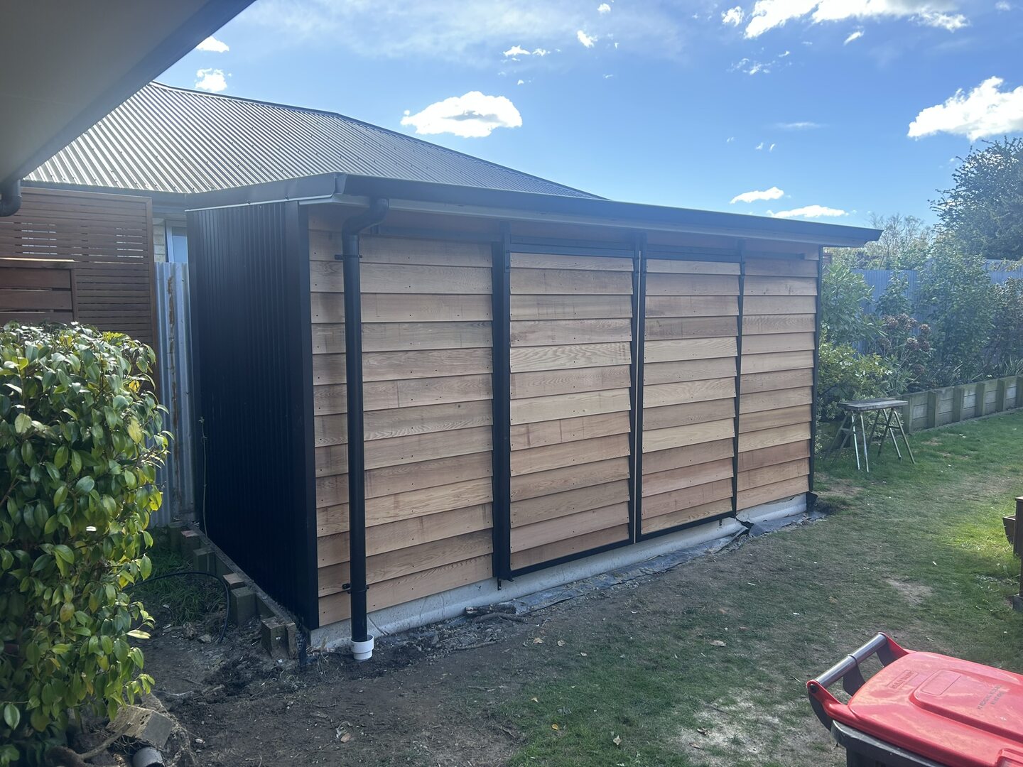 Cedar weatherboard garden store with black steel corner trims, a hipped corrugated roof and black spouting and downpipes, sitting on a concrete strip footing