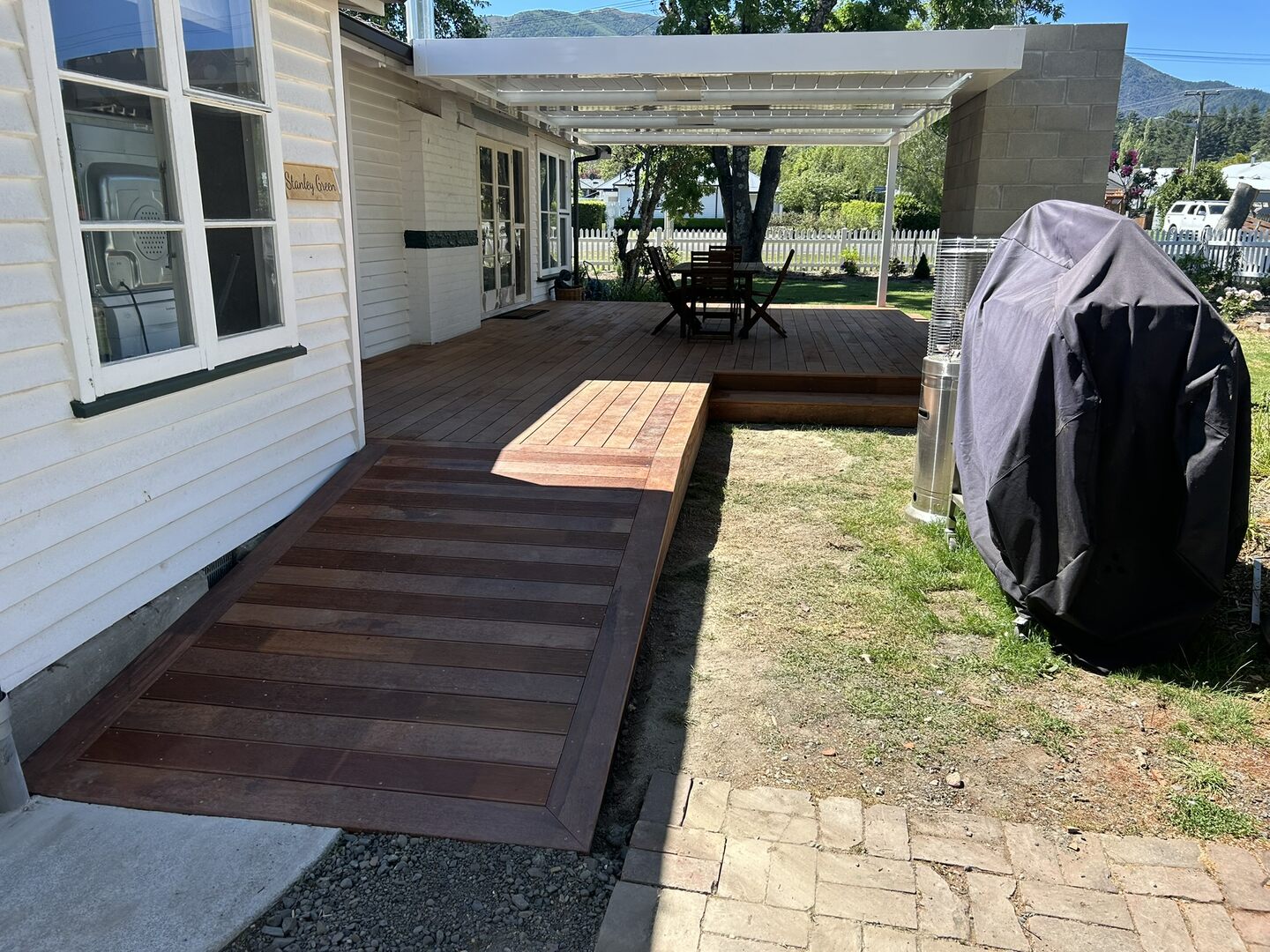Hardwood walkway and shallow ramp beside a weatherboard cottage, leading up to a raised deck under a white powder-coated pergola