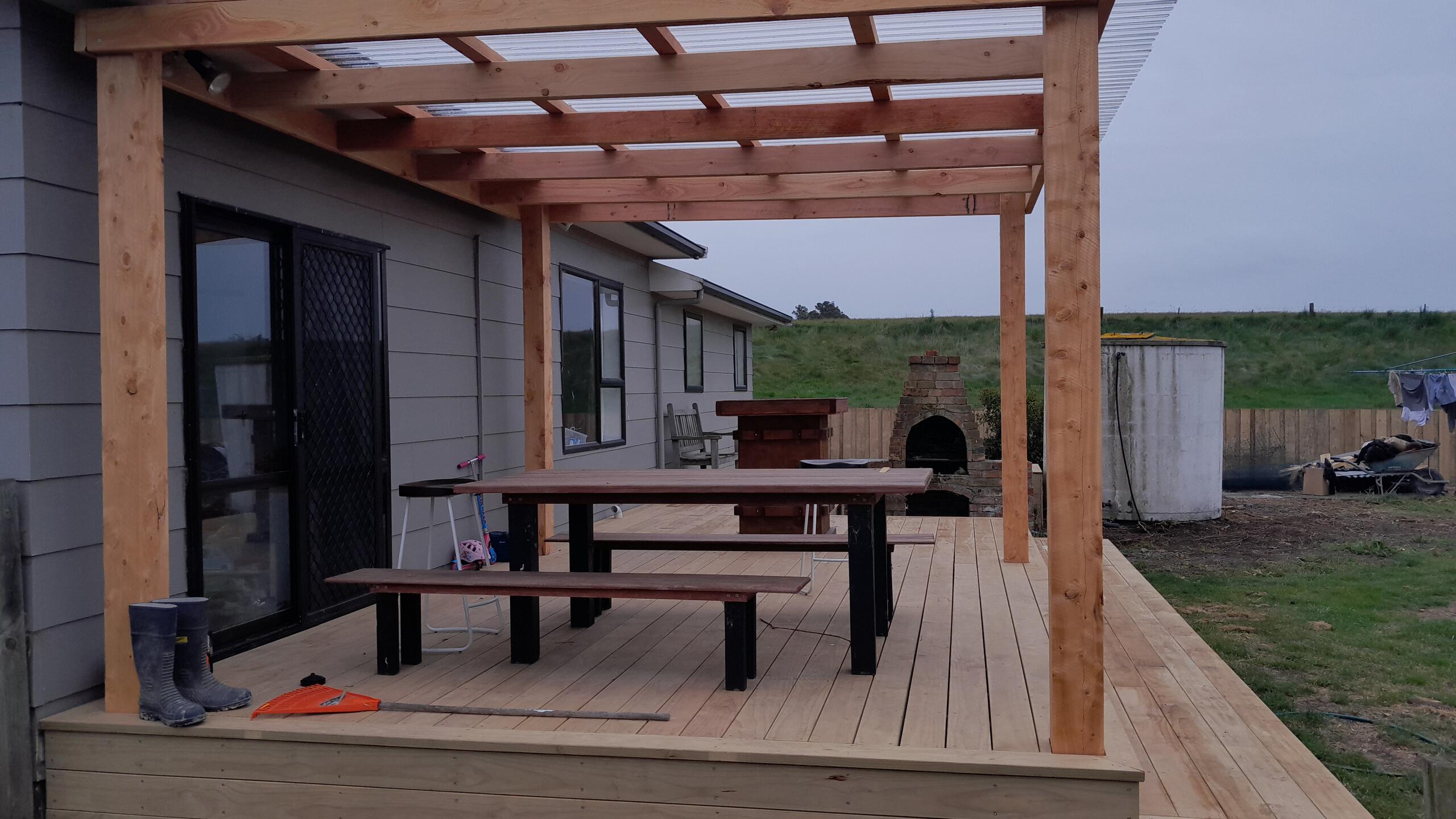 Cedar post-and-beam pergola with clear roof sheeting over a new timber deck, with a brick outdoor fireplace and a paddock beyond
