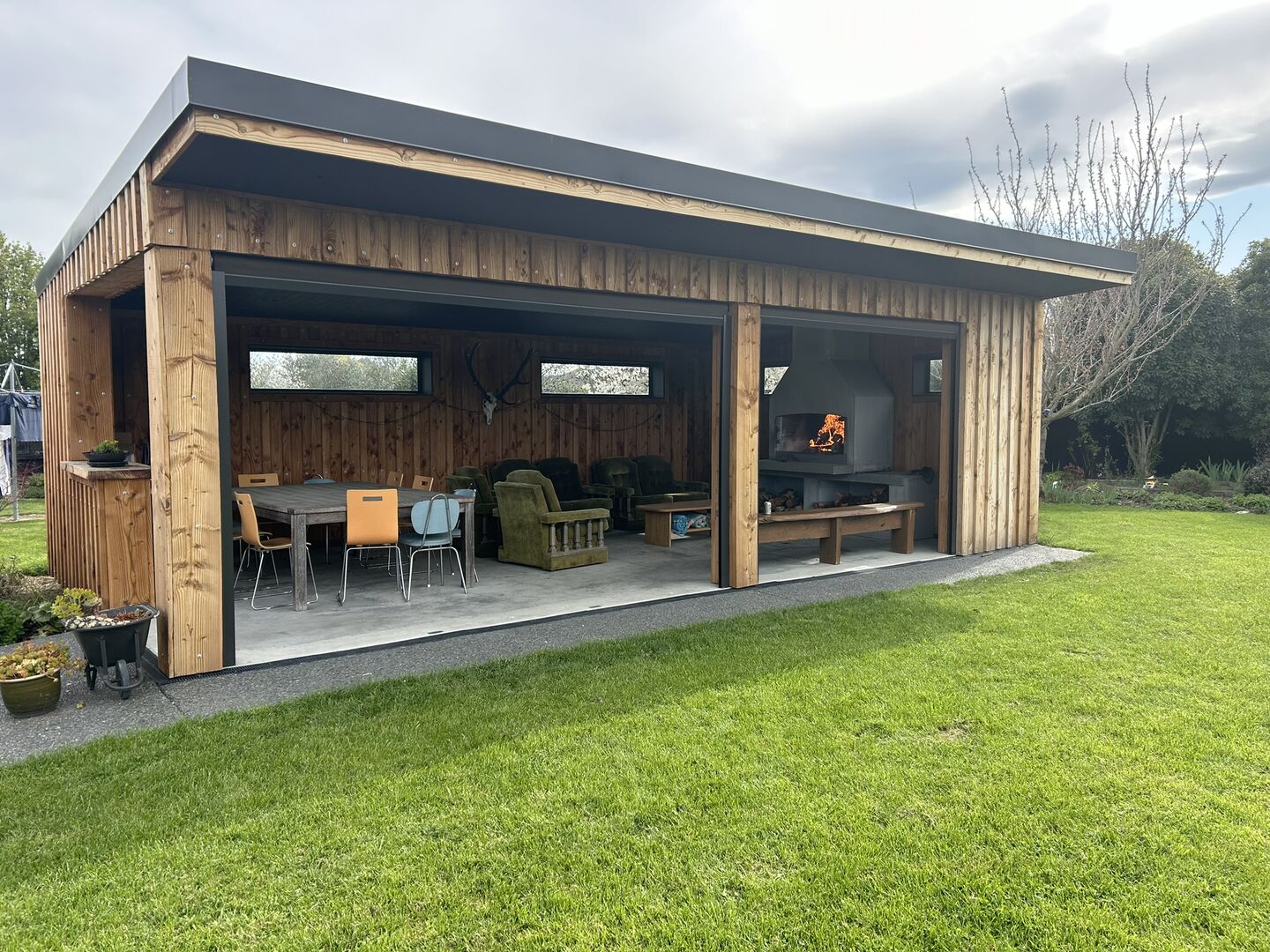 Cedar-clad outdoor pavilion with a flat dark roof, open on two sides, seen across the lawn with a dining table, armchairs and a fireplace inside