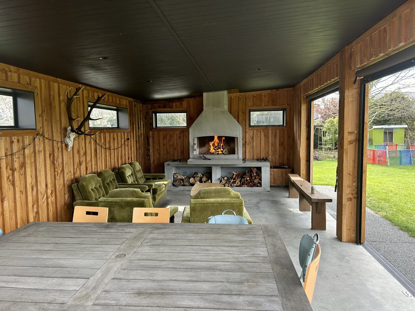 Inside the pavilion, looking at the plastered outdoor fireplace with a fire lit and firewood stacked in the recesses below, with vertical cedar lining and a dark-stained ceiling