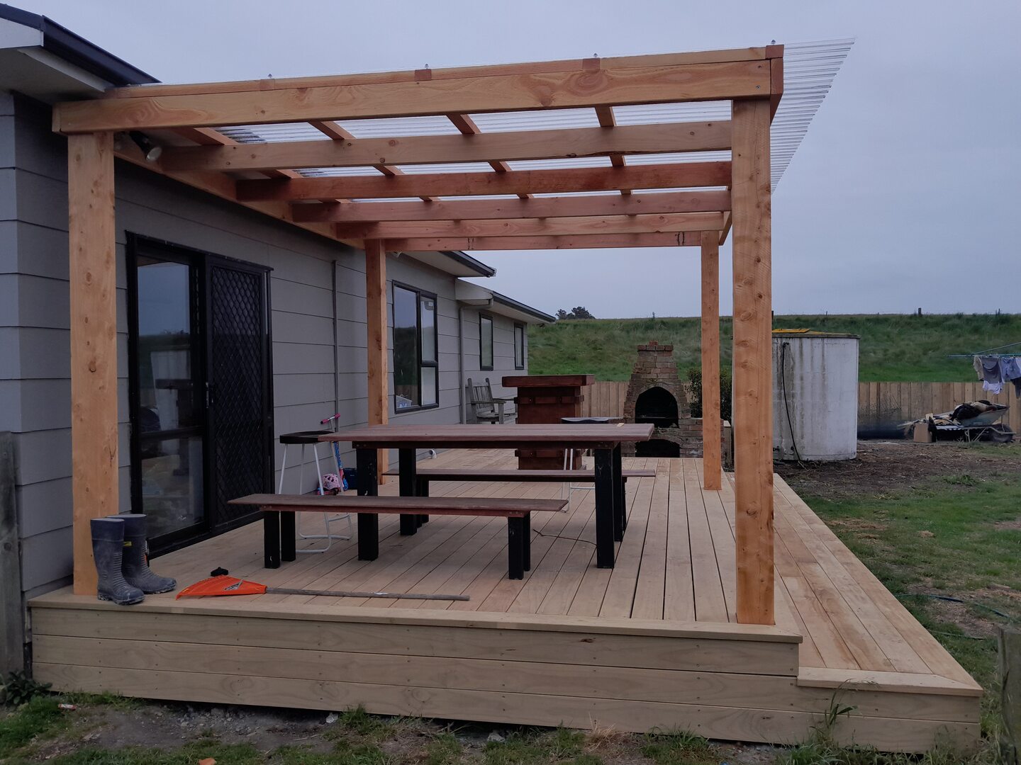Cedar post-and-beam pergola with clear roof sheeting over a new timber deck, with a hardwood table and benches, a brick outdoor fireplace and a paddock beyond