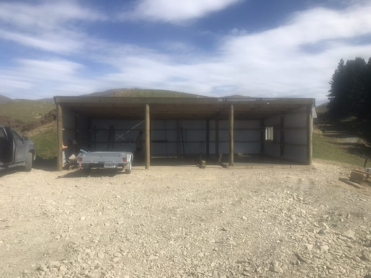 Pole shed with three open bays, round timber posts and profiled steel cladding, on a gravel pad below tussock hills