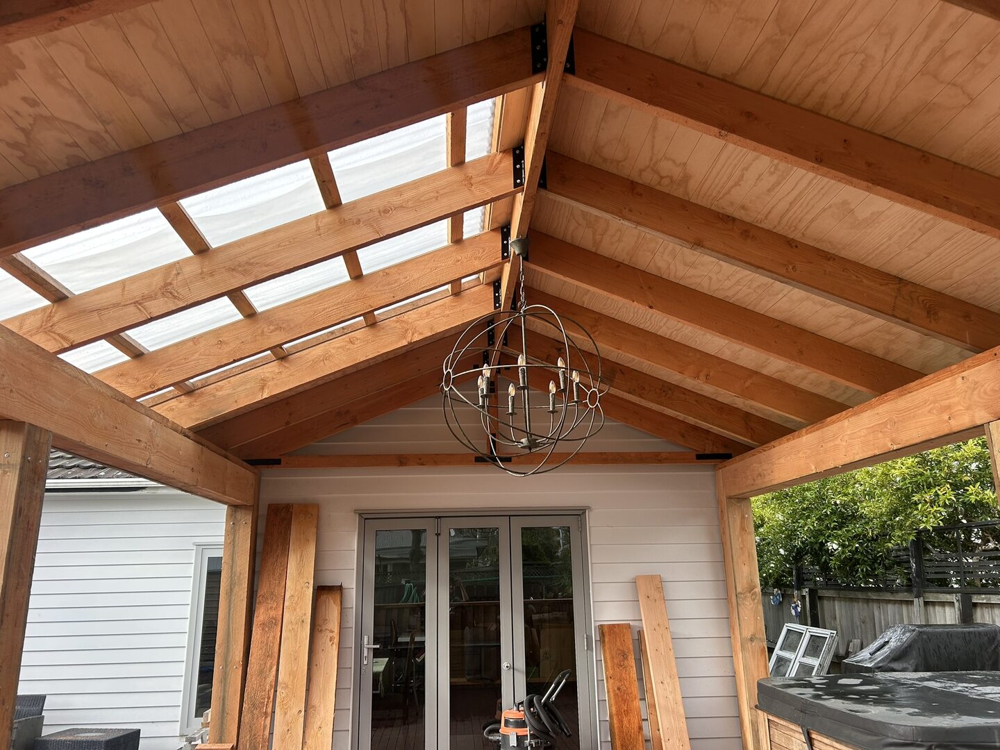 The gable roof of the outdoor room seen from below, with exposed timber rafters, a lined plywood ceiling, clear roof sheeting to one pitch and black steel brackets at the ridge
