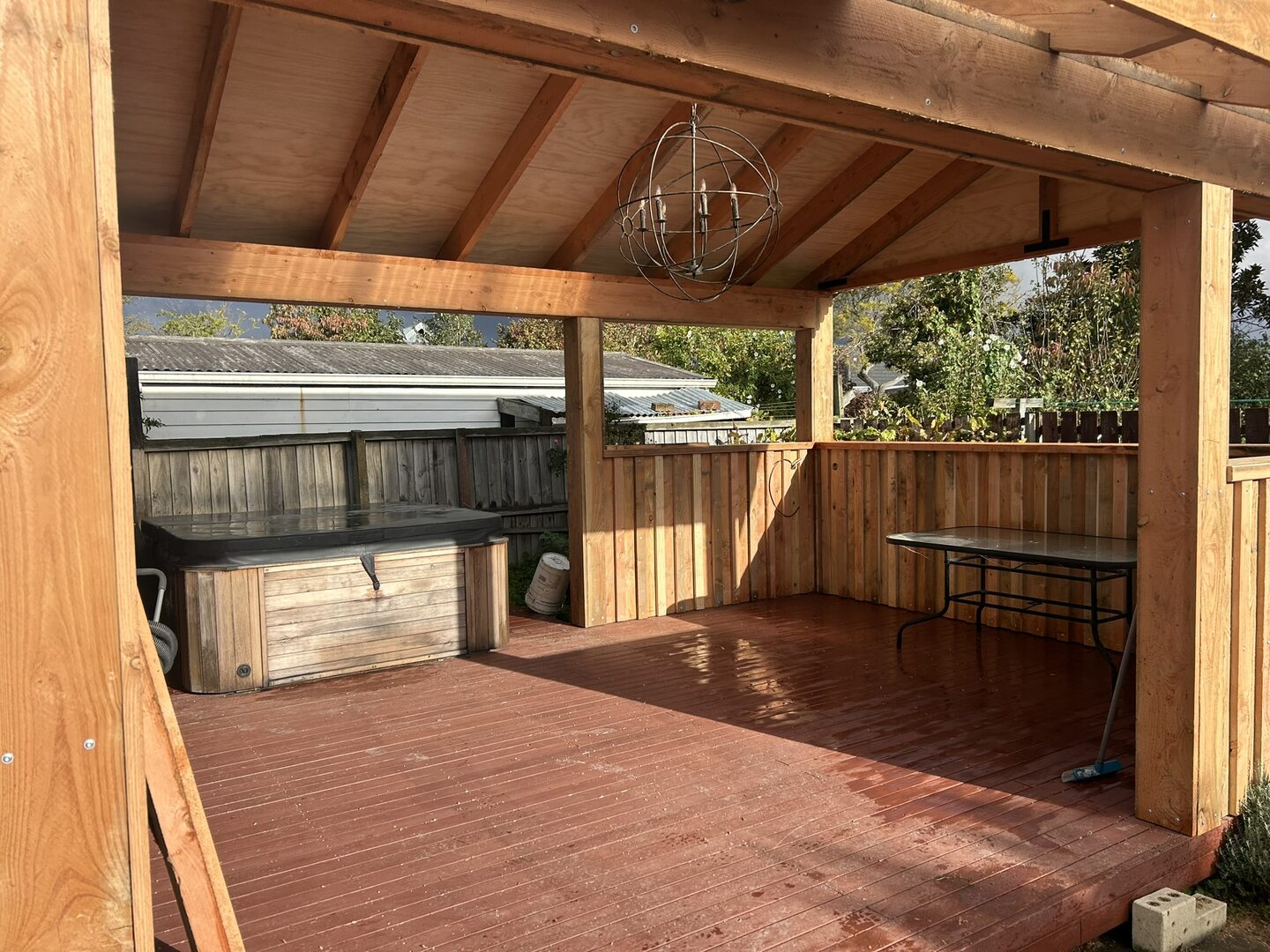 Inside the covered deck, looking across stained decking to the spa pool, with cedar balustrade infill panels and a round pendant light hung from the rafters